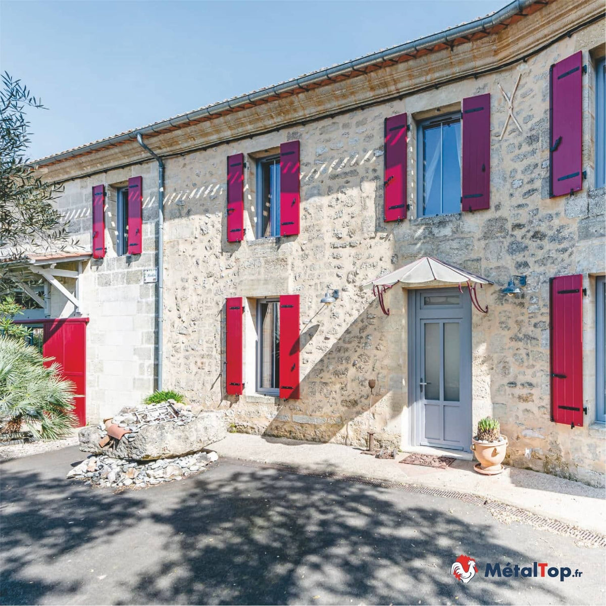 Façade en pierre avec volets rouges et fuchsia, porte d'entrée grise et auvent blanc