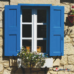 Fenêtre blanche à carreaux, volets bleus et jardinière fleurie blanche sur un mur en pierre