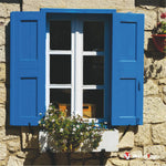 Fenêtre blanche aux volets bleus et jardinière fleurie sur un mur en pierre