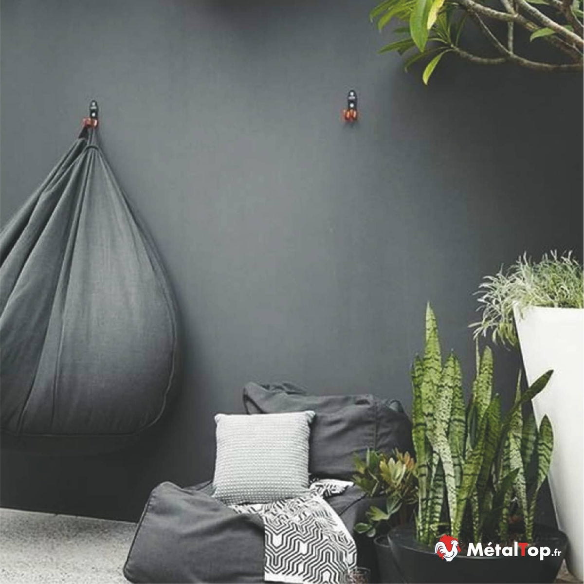 Crochets muraux noirs supportant un pouf suspendu gris, coussins et plantes vertes