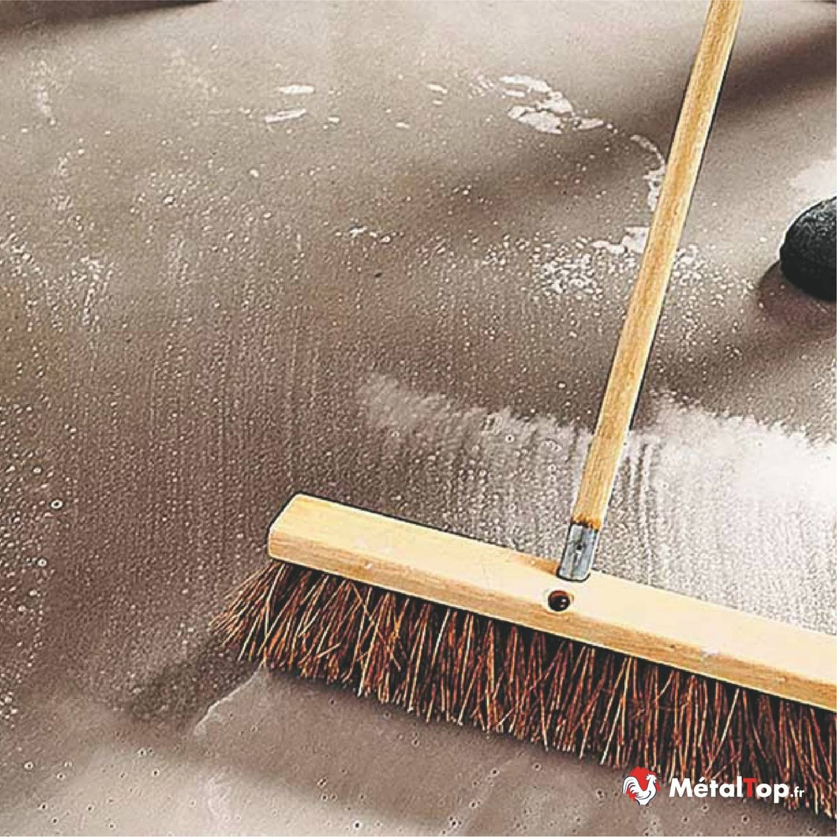 Balai-brosse à manche en bois balayant un sol gris humide couvert de mousse blanche