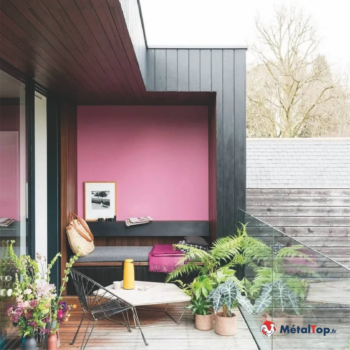 Terrasse en bois avec mur rose, banc gris, fauteuil filaire et plantes vertes