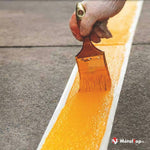 Application d'une peinture orange sur une bande blanche tracée au sol en béton avec un pinceau
