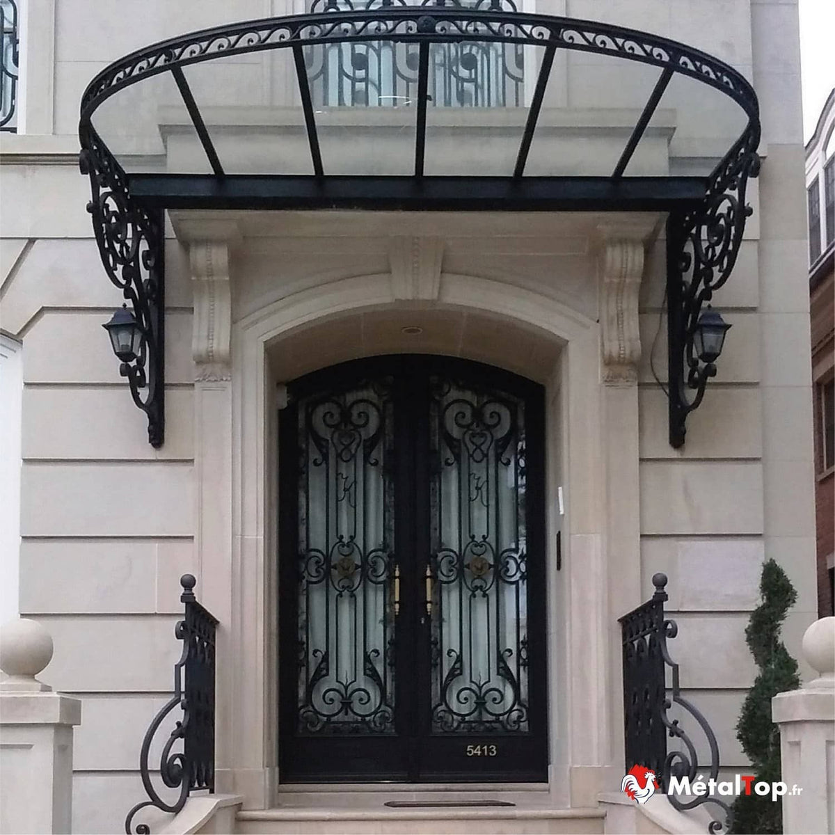 Porte cintrée en fer forgé noir sous auvent en verre à ferronnerie, façade en pierre claire