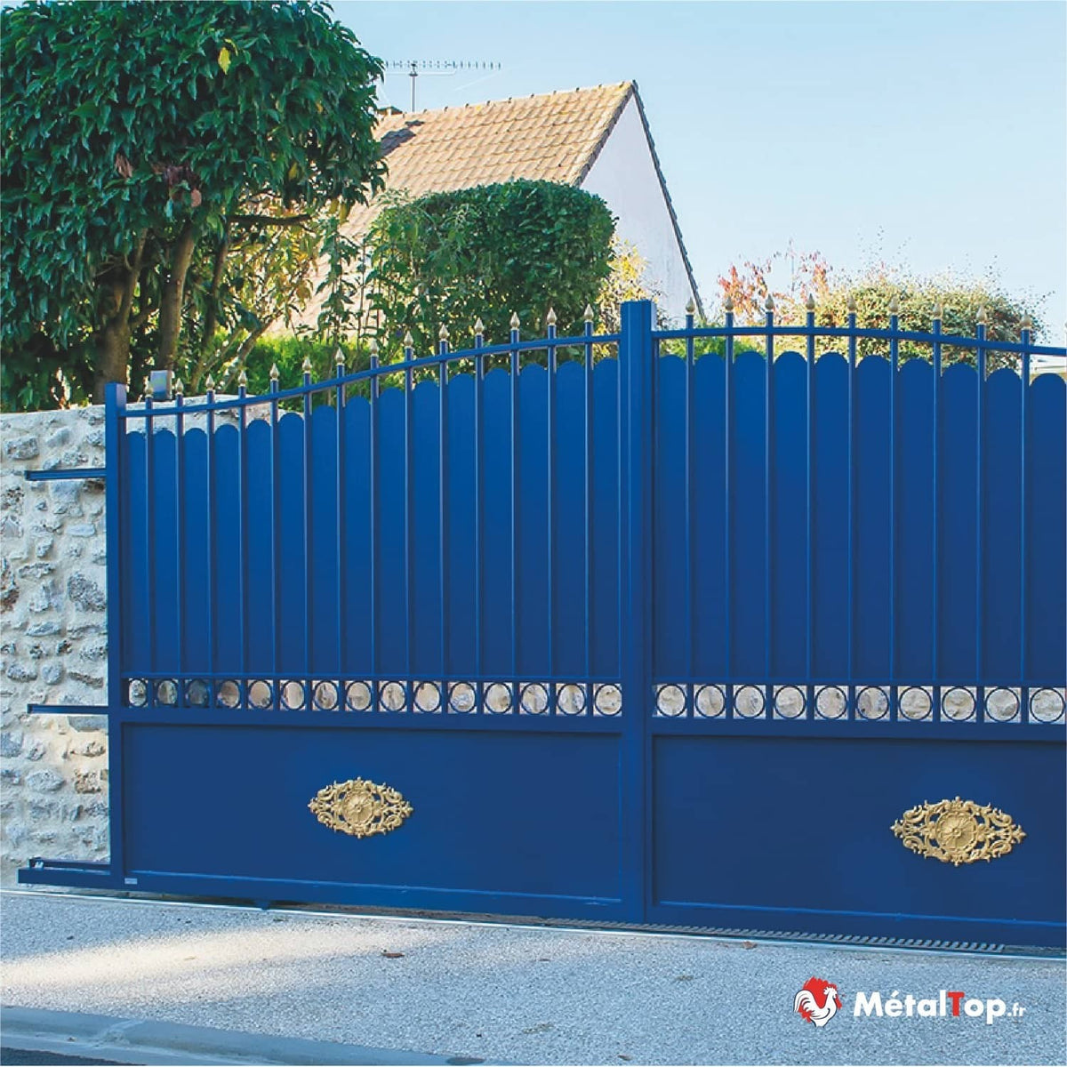 Portail coulissant bleu en métal avec ornements dorés et pointes décoratives, devant un mur en pierre