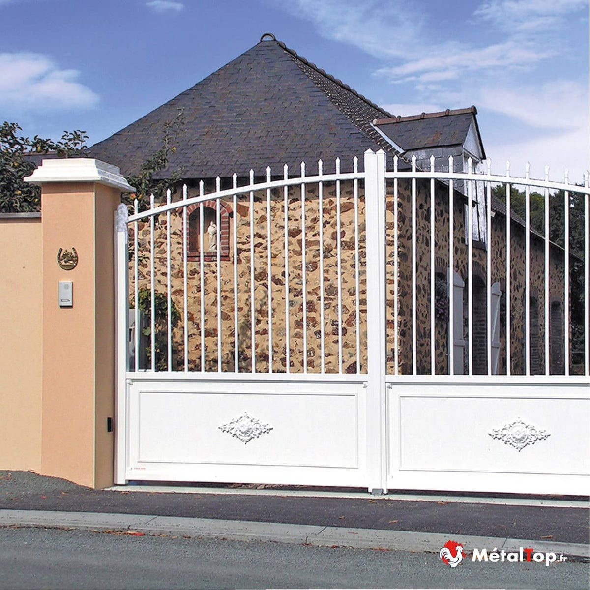 Portail blanc à barreaux et motifs décoratifs, devant une maison en pierre