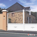 Portail blanc à barreaux et motifs décoratifs, devant une maison en pierre