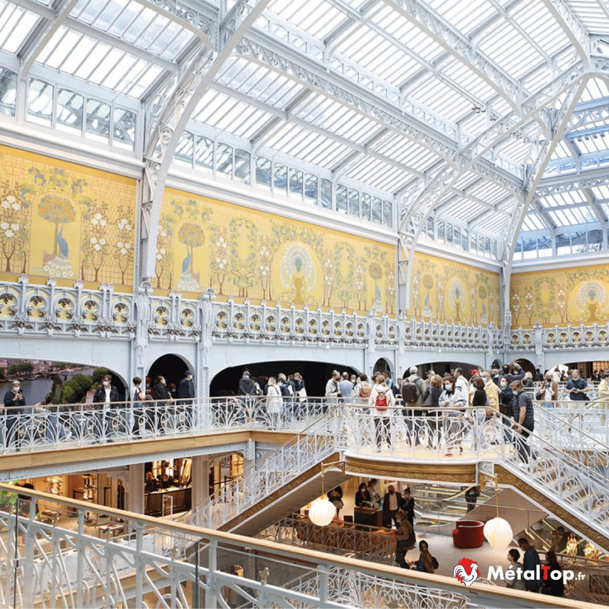 Verrière et ferronnerie blanches, fresques jaunes aux paons et visiteurs dans un grand magasin