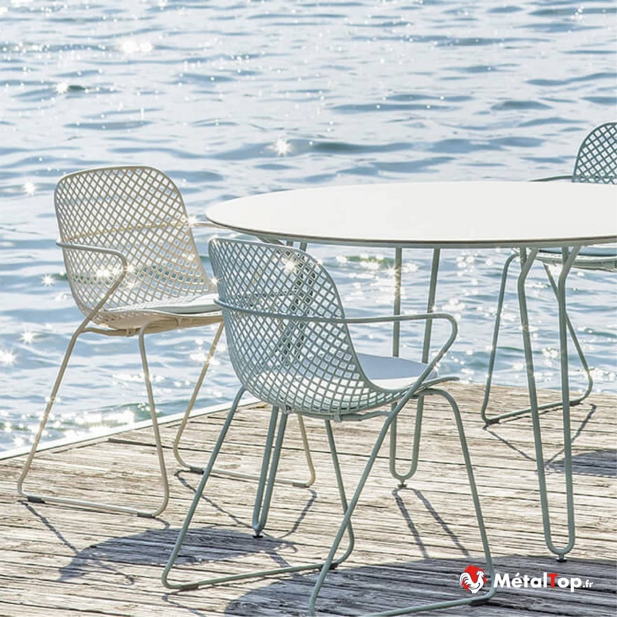 Chaises en métal ajourées et table ronde blanche sur une terrasse en bois au bord de l'eau