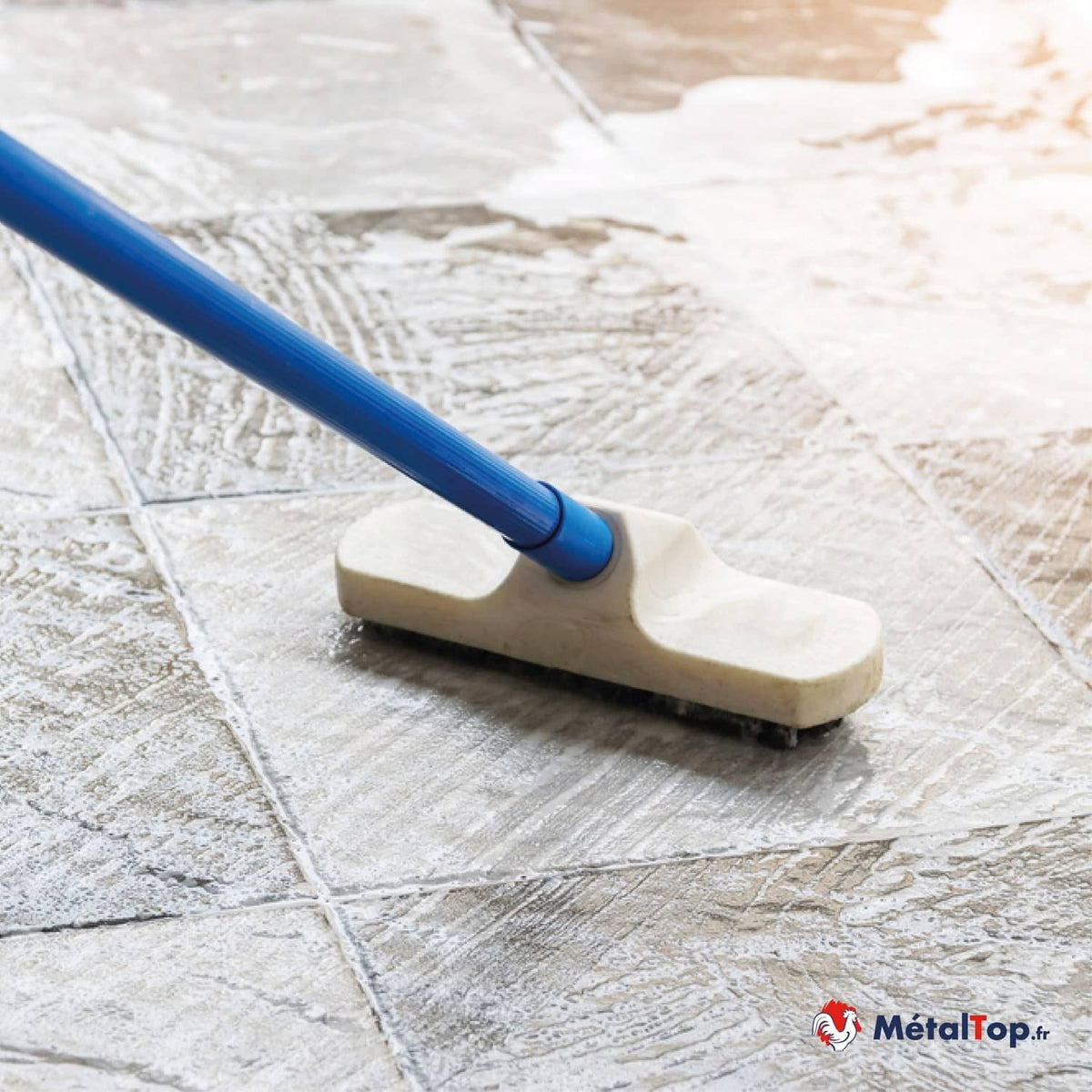 Brosse à manche bleu nettoyant un carrelage beige texturé couvert de mousse blanche