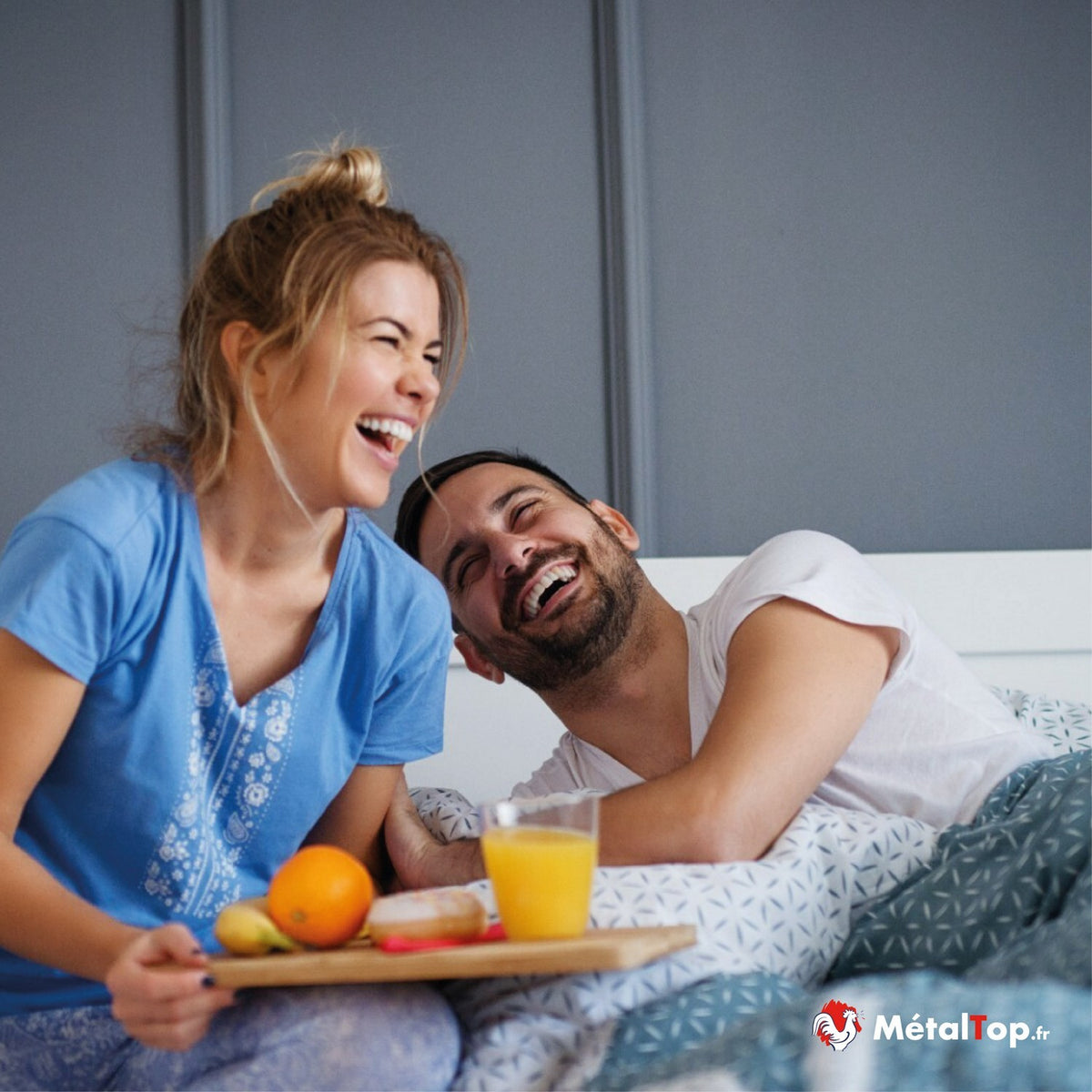 Couple riant au lit, la femme tient un plateau petit-déjeuner avec jus d'orange et fruits