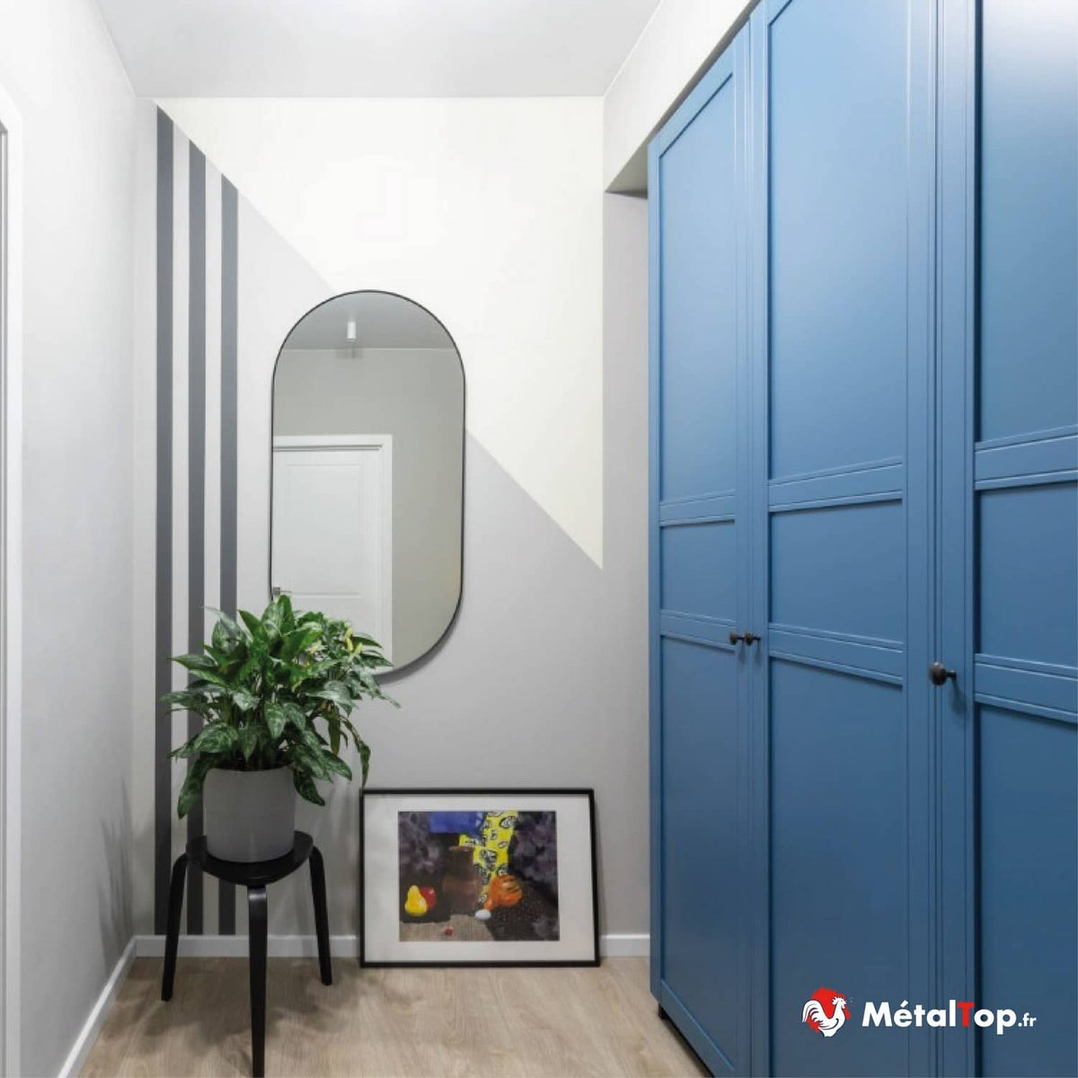 Couloir avec armoire bleue, miroir ovale, plante verte sur tabouret noir et tableau au sol