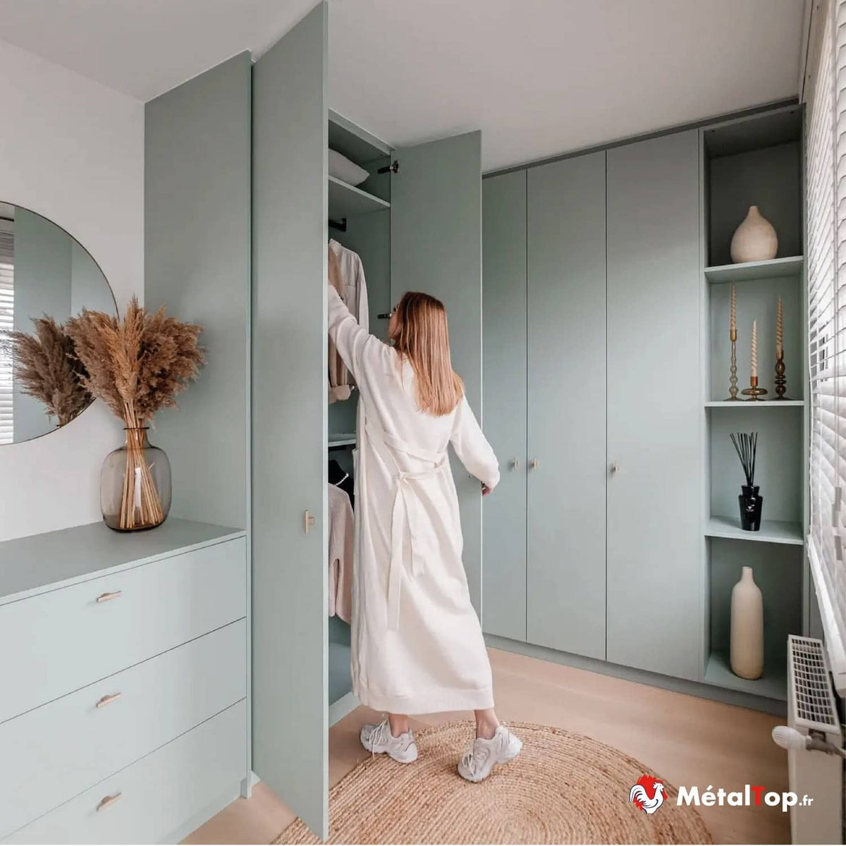 Femme en peignoir blanc rangeant ses vêtements dans un dressing vert sauge avec étagères