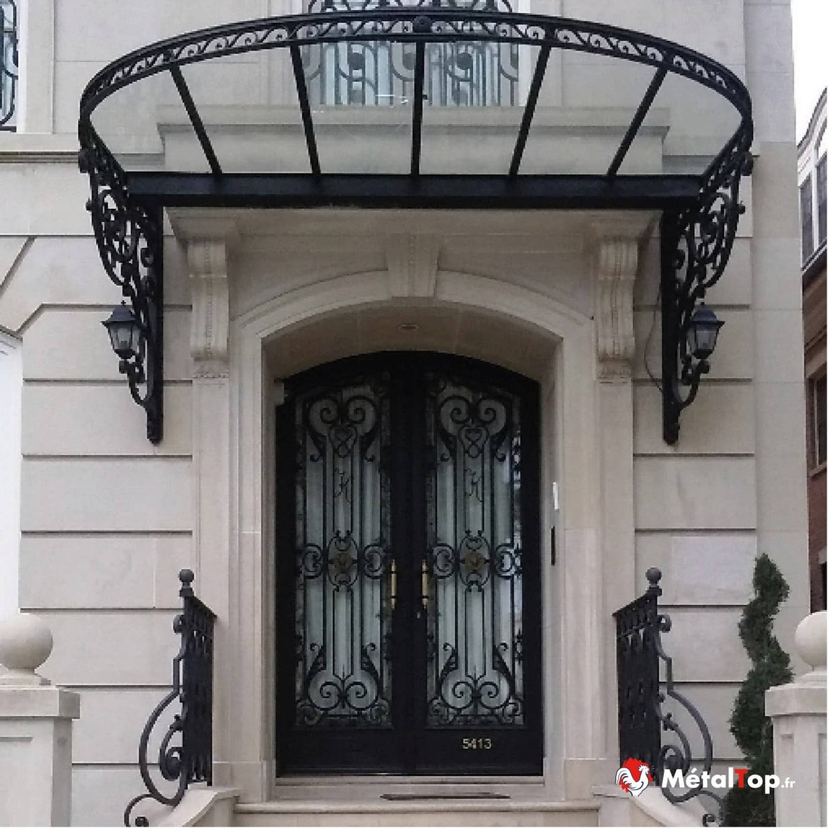 Auvent en verre et fer forgé noir au-dessus d'une double porte d'entrée ornée de ferronnerie